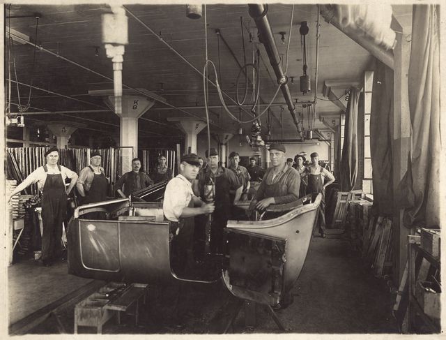 Workers in a factory assembling a car body. Some are standing around, others are working on the vehicle