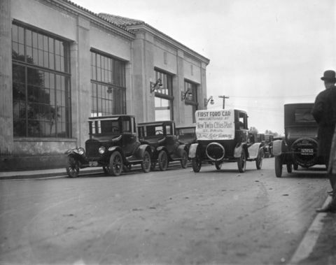 1909 Ford Model T - Several Ford Model T cars are parked in front of a building, one of which carries a sign indicating it is the first Ford car manufactured a…