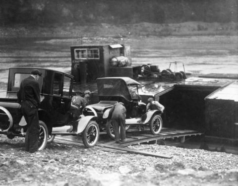 1909 Ford Model T - Workers are seen loading early automobiles onto a transport platform, possibly a ferry, with a building and machinery in the background.