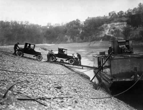 1909 Ford Model T - Men push two Ford Model T cars onto a barge next to a riverbank with a building on a hill in the background.