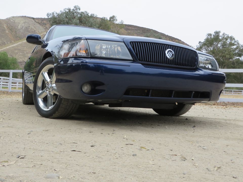 Front view of a dark blue 2003 Mercury Marauder parked on a dirt road with a white fence and hills in the background
