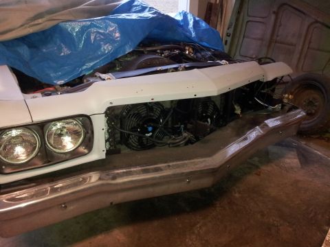 1976 Ford Ranchero - A white vehicle's front end is partially covered by a blue tarp, revealing dual headlights and an engine bay with cooling fans.