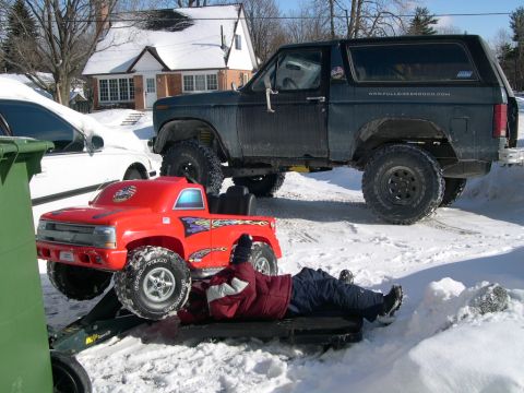 1986 Ford Bronco - A person works on a red toy truck while a lifted 1986 Ford Bronco sits nearby in a snowy setting.