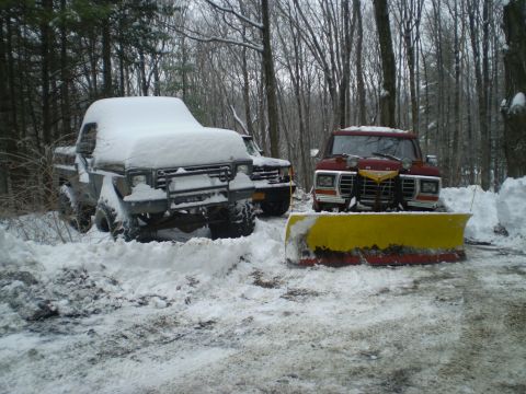 1979 Ford Bronco - Two trucks, one a 1979 Ford Bronco with a snow plow, are parked in a snowy wooded area.