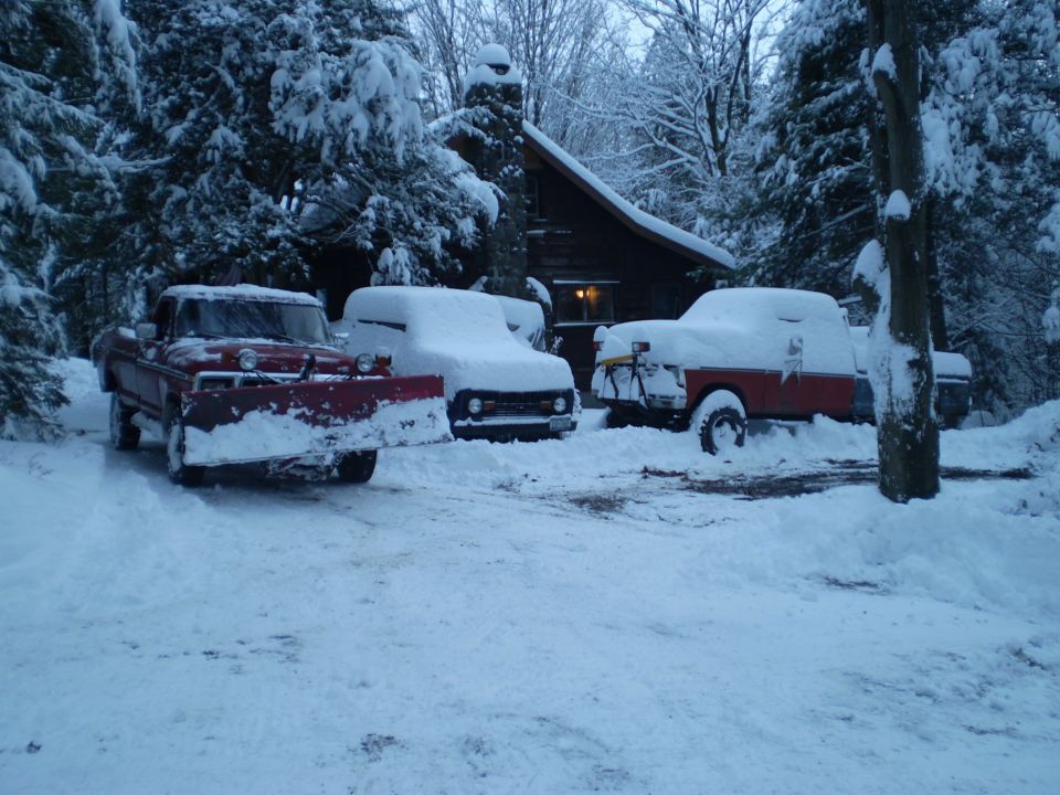 Three snow-covered trucks parked in front of a cabin, one with a plow