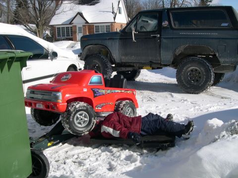 1986 Ford Bronco - A dark green Ford Bronco is parked in the snow, with a person lying on a sled underneath a red toy truck.