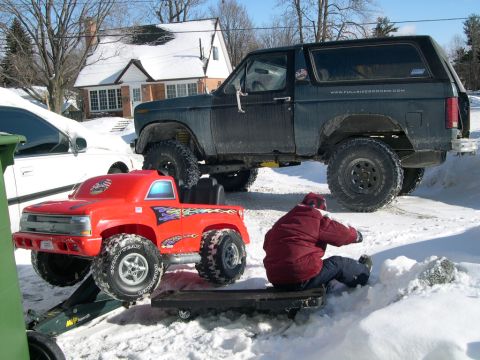 1986 Ford Bronco - A dark 1986 Ford Bronco is parked in the snow next to a red toy truck, with a person sitting on a sled nearby.