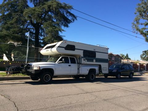 1986 Yugo GV - A white pickup truck with a large camper shell is parked on the street, towing a dark SUV behind it.