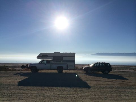 1986 Yugo GV - A truck camper and a dark SUV are parked on a dirt road under a bright sun, with a layer of fog in the distance.