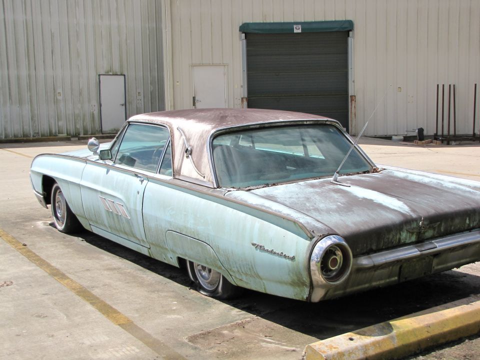 Old light blue car with a rusty roof parked outside a warehouse