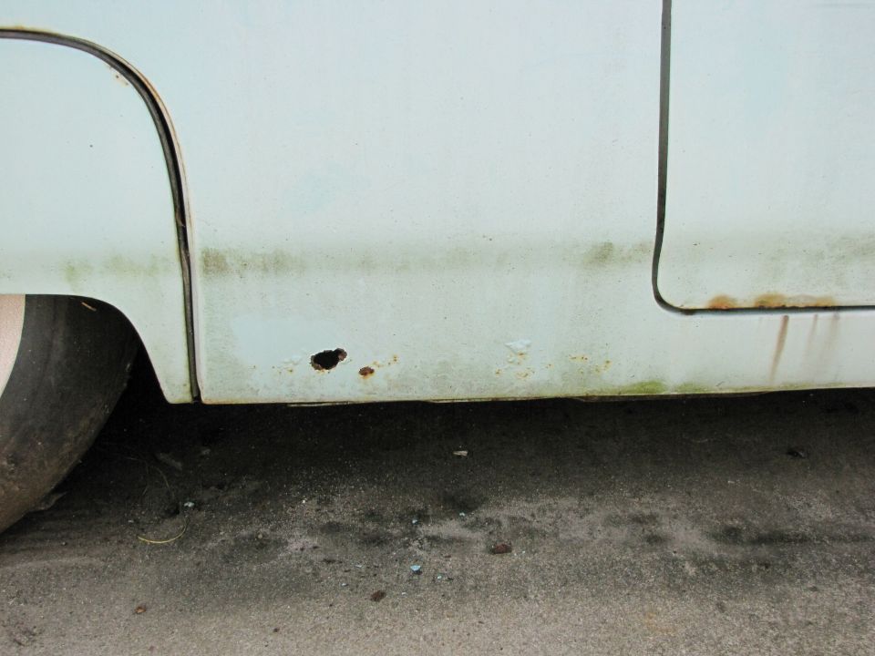 Close-up of a light blue car's lower door panel showing rust and a hole. A tire is partially visible on the left