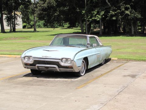 1963 Ford Thunderbird - A light blue 1963 Ford Thunderbird is seen parked in a lot with trees in the background.