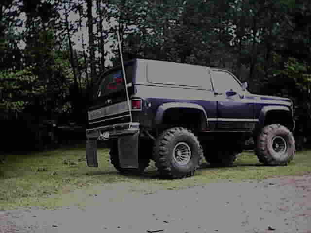 Lifted purple two-door Blazer SUV on large mud tires, rear 3/4 view in wooded clearing