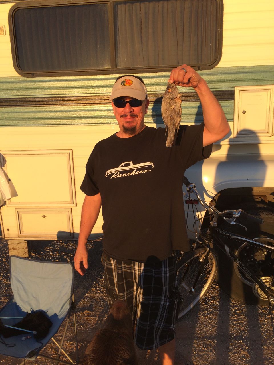 Man in a black t-shirt with a Ranchero logo holds up a small fish in front of an RV