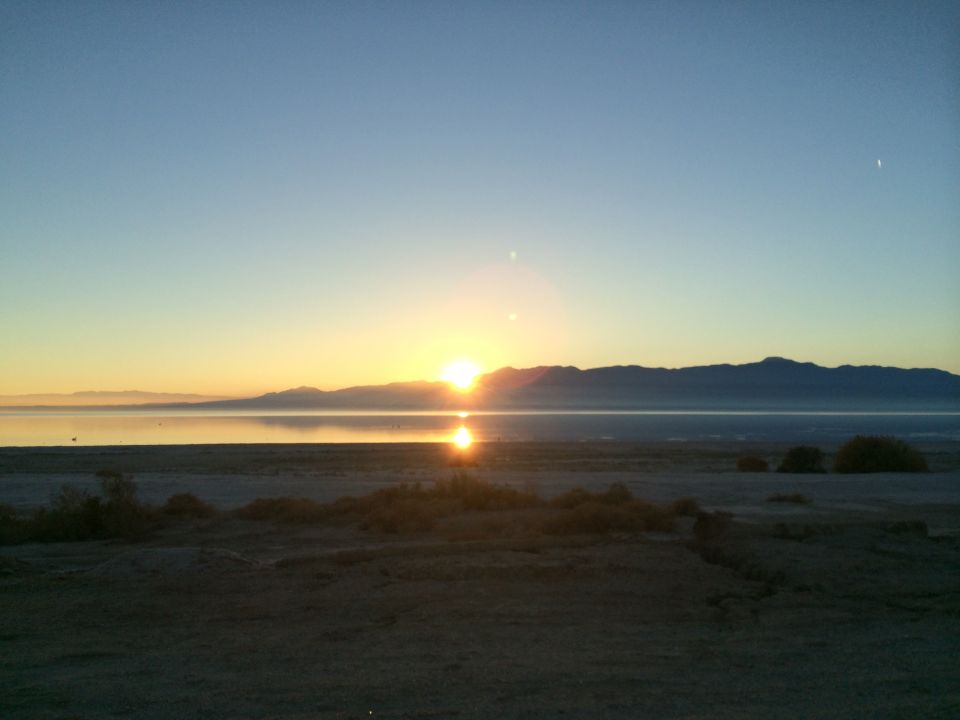 Sunset over a calm lake with distant mountains and a sandy shore in the foreground