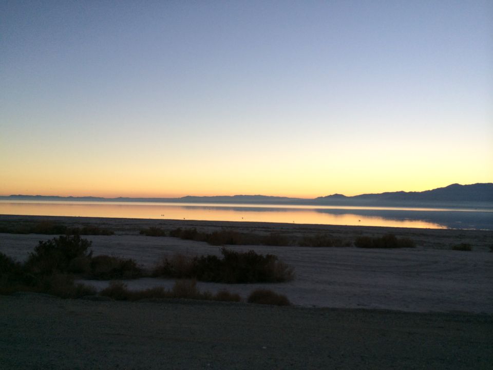 Desert landscape with mountains in the distance and a calm body of water reflecting the sunset colors