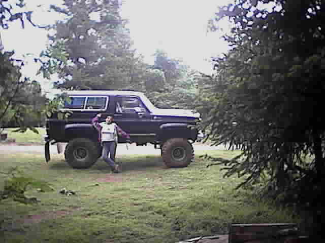 Lifted dark Chevrolet Blazer on large off-road tires with a person standing in front, in a grassy wooded yard.