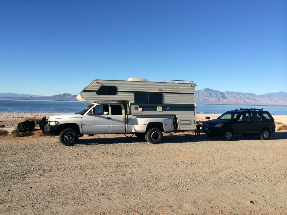 White pickup truck with camper shell towing a black SUV near a body of water