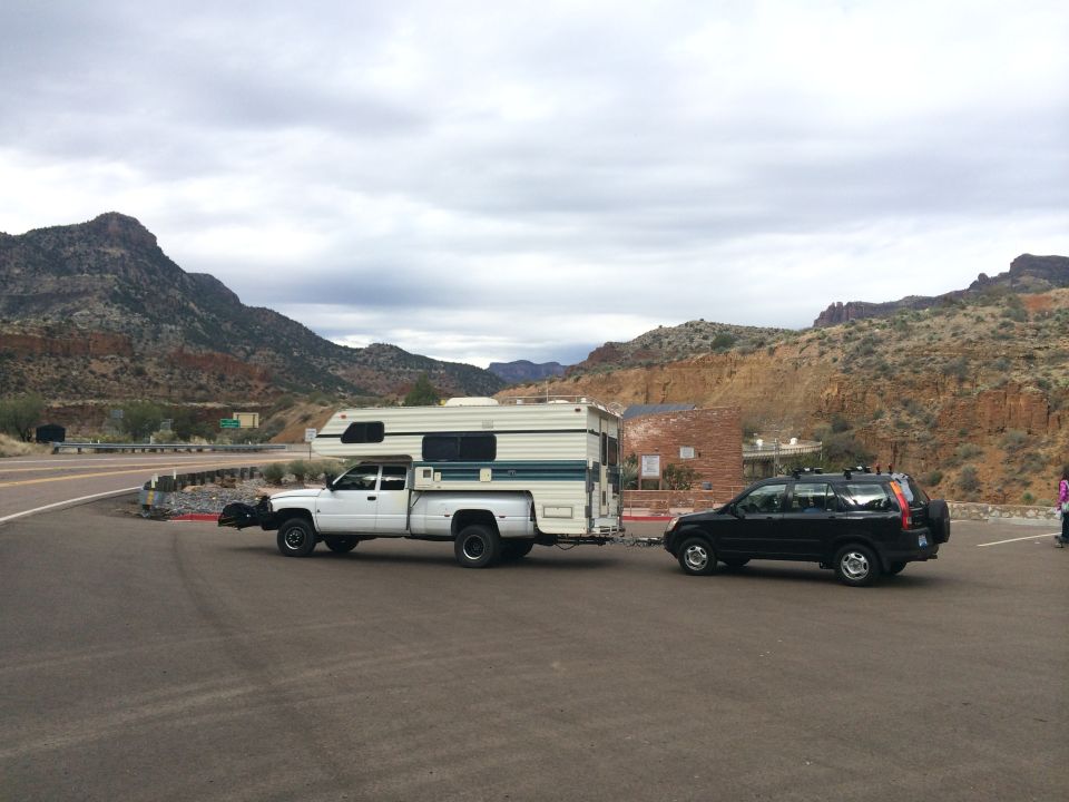 White pickup truck with camper shell towing a black SUV on a road with mountains in the background