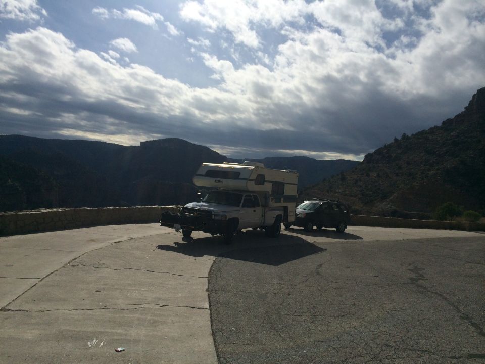 Truck with camper shell and an SUV parked on a paved overlook with mountains in the background