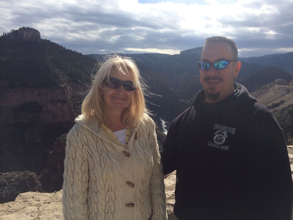 Woman in a cream cable-knit cardigan and man in a black hoodie stand on a rocky outcrop with a canyon vista behind them