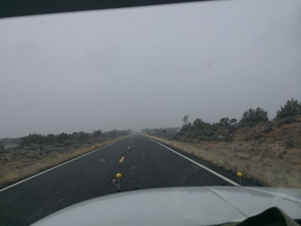 View of a two-lane road disappearing into fog, with scrub brush and hills on either side. The hood of a vehicle is visible in the foreground