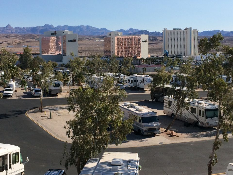 RV park scene with multiple recreational vehicles parked, hotels in the distance under a clear blue sky