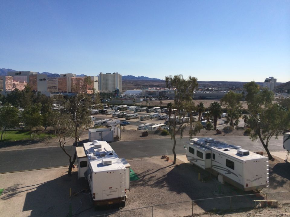 RV park scene with multiple recreational vehicles parked, buildings and mountains in the distance under a clear blue sky