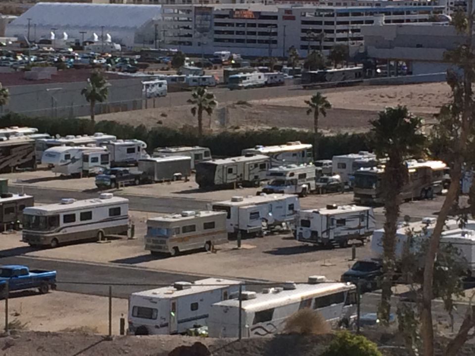 Many RVs and trailers parked in an outdoor lot with palm trees and buildings in the background