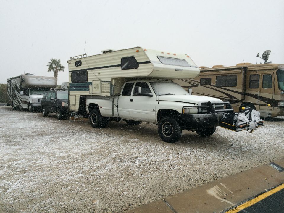 White pickup truck with a camper shell parked in snow, next to an RV and another vehicle