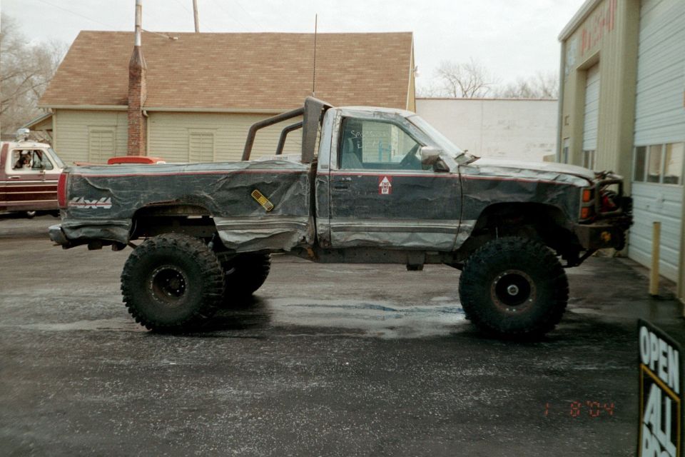 Modified pickup truck with large tires and roll bar, parked next to a building. Another pickup truck is visible to the left