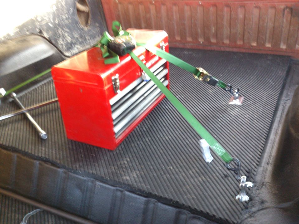 Red toolbox secured with green straps in a truck bed with a spare tire nearby