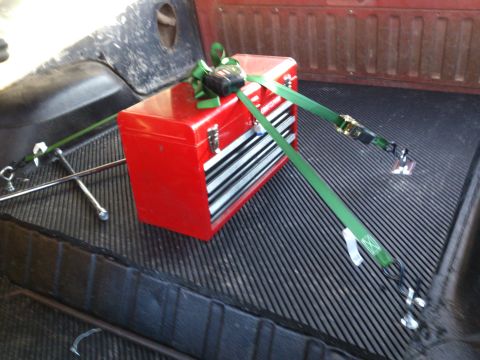1986 Ford Bronco - A red toolbox is strapped down in the bed of a pickup truck, alongside a jack and tire iron.