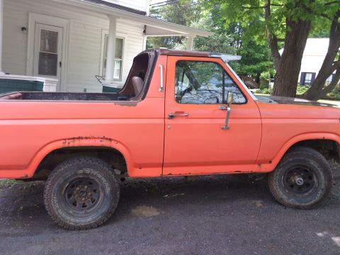 1986 Ford Bronco - This orange SUV is parked in front of a house with a porch and trees in the background.