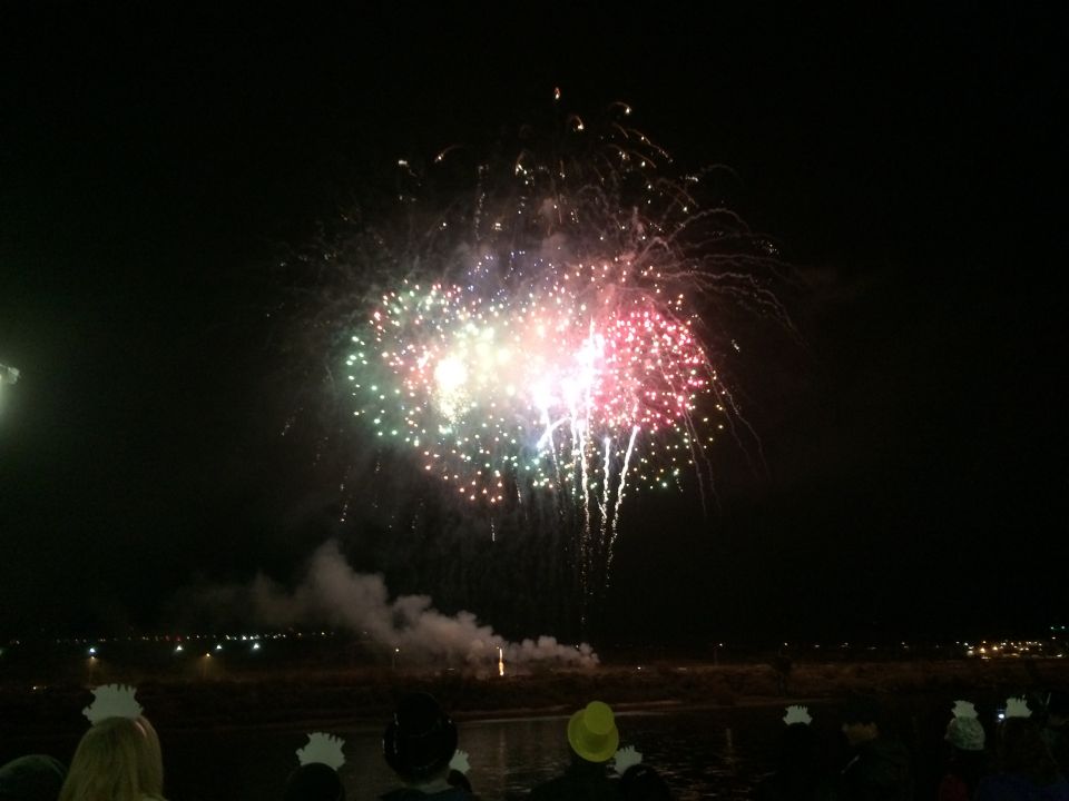 Fireworks explode in the night sky over a river as people watch from the shore