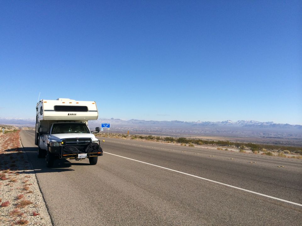 White pickup truck with camper shell on highway, distant mountains under clear blue sky