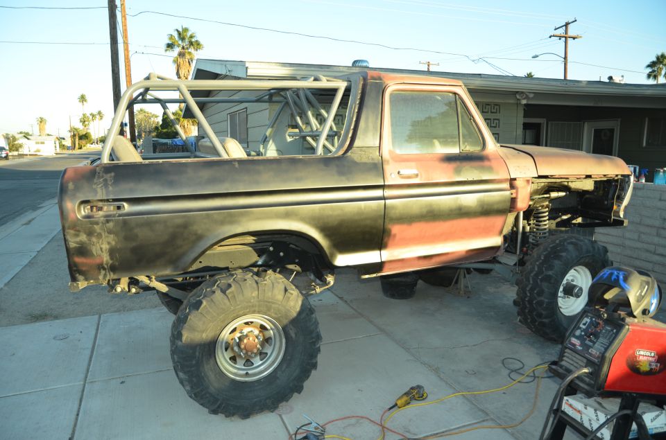 Ford Bronco body with roll cage, missing front end, on large tires. Welding equipment nearby