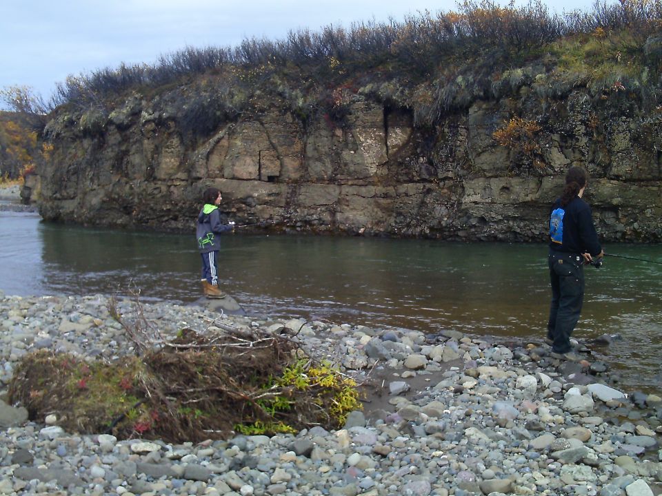 Two people fishing in a river with a rocky bank and a cliff face behind them