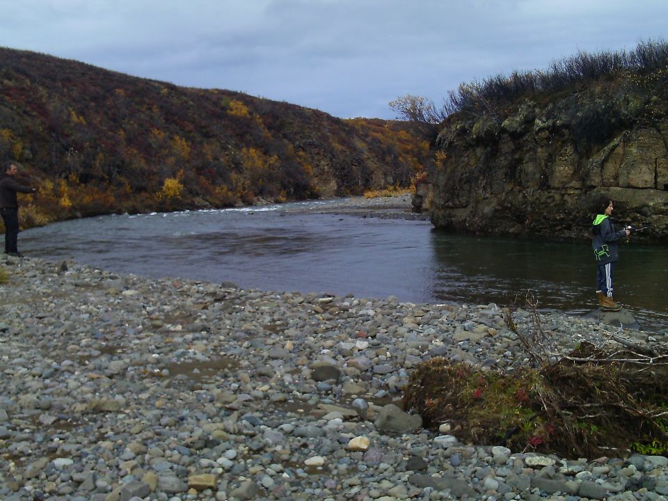 Rocky riverbank with two people fishing, autumn foliage on hillsides under a cloudy sky