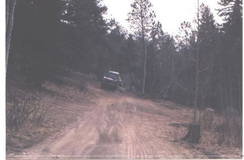 1996 Ford Bronco - A dark-colored SUV navigates a dirt road through a wooded area.