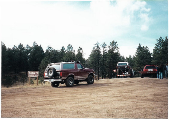 Maroon 1996 Ford Bronco parked on dirt road, with a trailer carrying another vehicle and a red car behind it