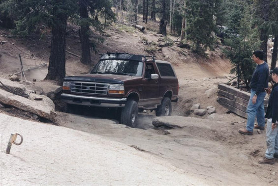 Maroon Ford Bronco navigating a rocky off-road trail with two people watching