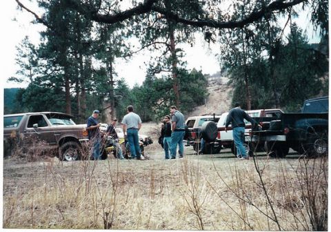 1996 Ford Bronco - A group of people gather around a brown Ford Bronco and other vehicles in a dry, wooded landscape.