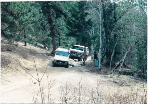 1996 Ford Bronco - Two SUVs, including a 1996 Ford Bronco, traverse a challenging off-road trail surrounded by trees.