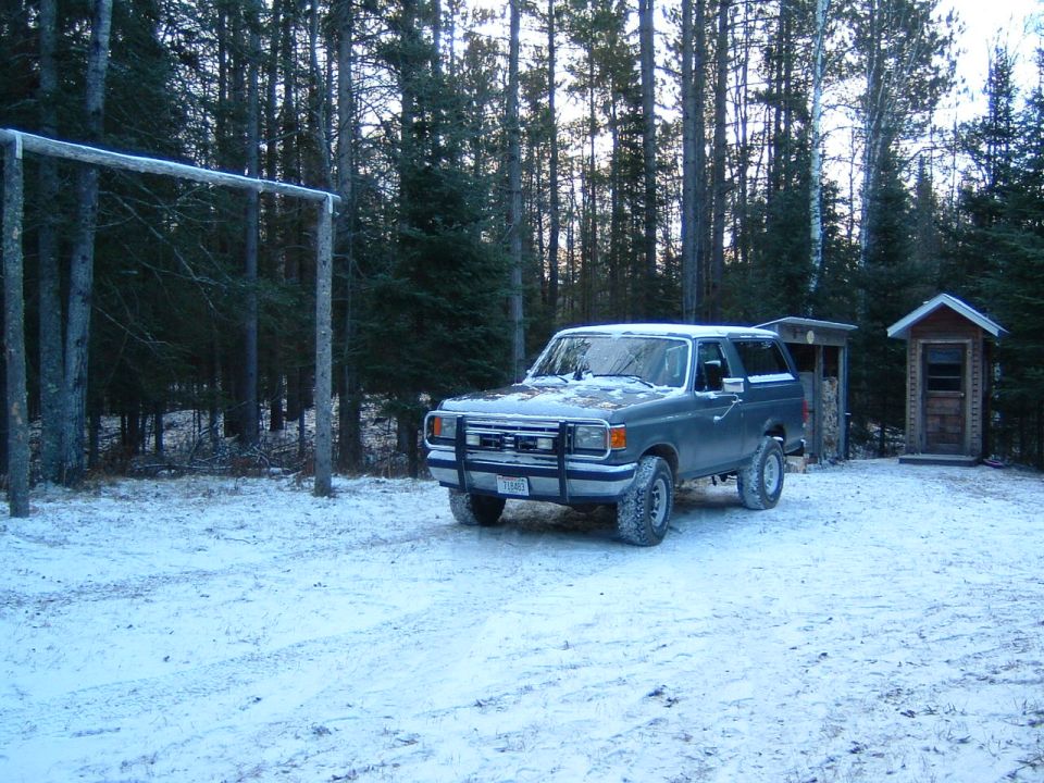 Gray Ford Bronco with brush guard on snowy clearing at dusk near small wooden shed