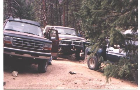 1996 Ford Bronco - Three off-road vehicles are parked on a dirt path surrounded by trees.