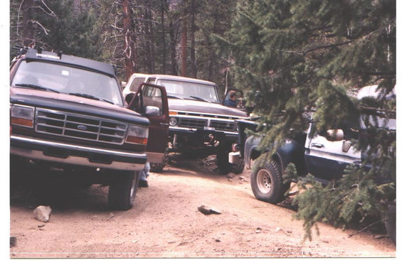 Dark blue Ford Bronco front-left view, parked on dirt road with trees