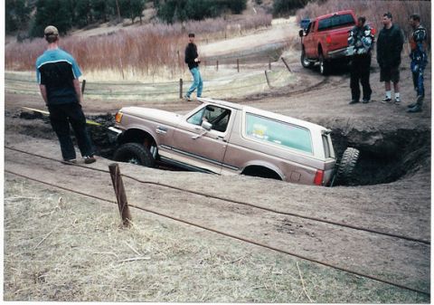 1996 Ford Bronco - A 1996 Ford Bronco is stuck in a hole while a red pickup truck and onlookers watch.