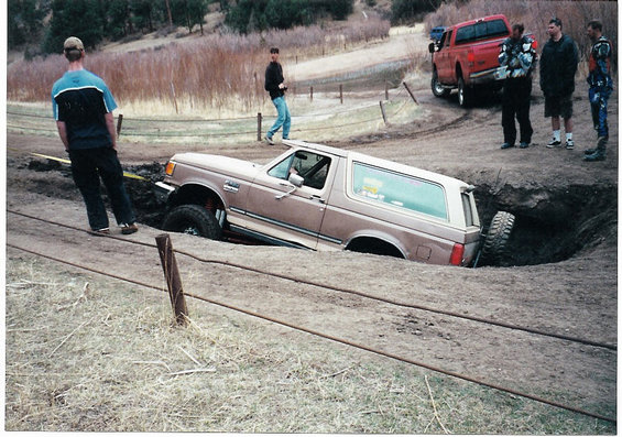 1996 Ford Bronco stuck in a deep dirt hole with a red pickup truck nearby