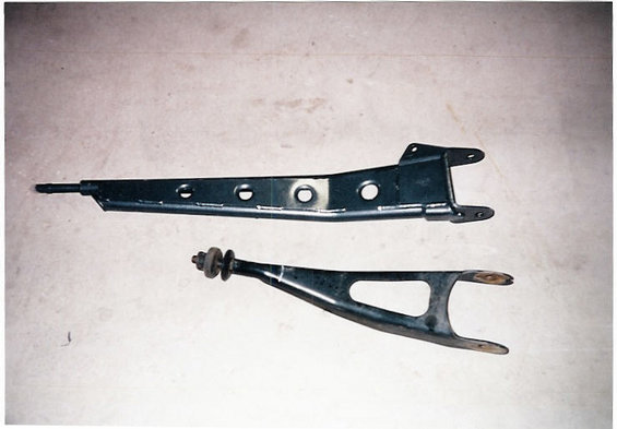 Two black suspension control arms on a light-colored surface. One is long and straight with circular cutouts, the other is curved with a rubber bushing